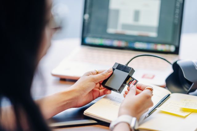 A person inserts an SD card into a reader next to a laptop on a desk