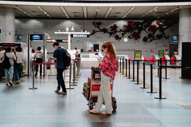 A traveler checks her phone while waiting with a luggage cart at a busy airport terminal