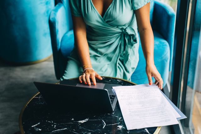 Adult woman in teal dress working on a laptop with documents, indoors