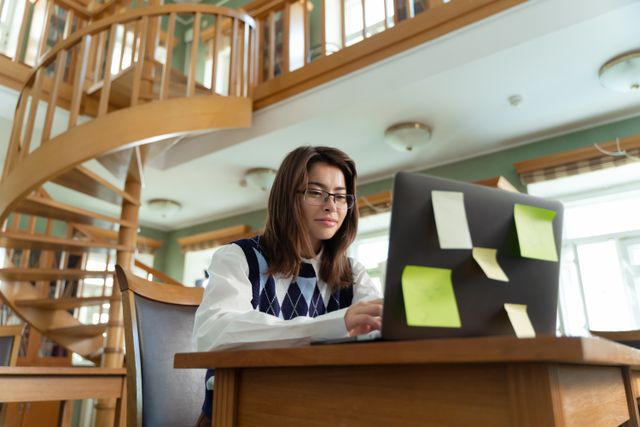 Focused young woman using laptop with notes in a bright room with spiral staircase